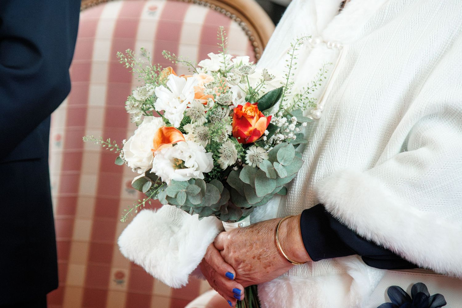 Bride in white fur wrap holding a wedding bouquet of white and orange roses with eucalyptus greenery.