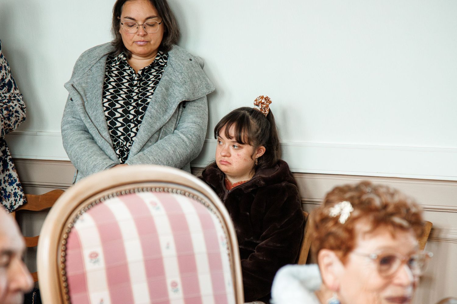 Woman and young girl seated at an elegant event, with ornate pink striped chairs in a formal interior setting.