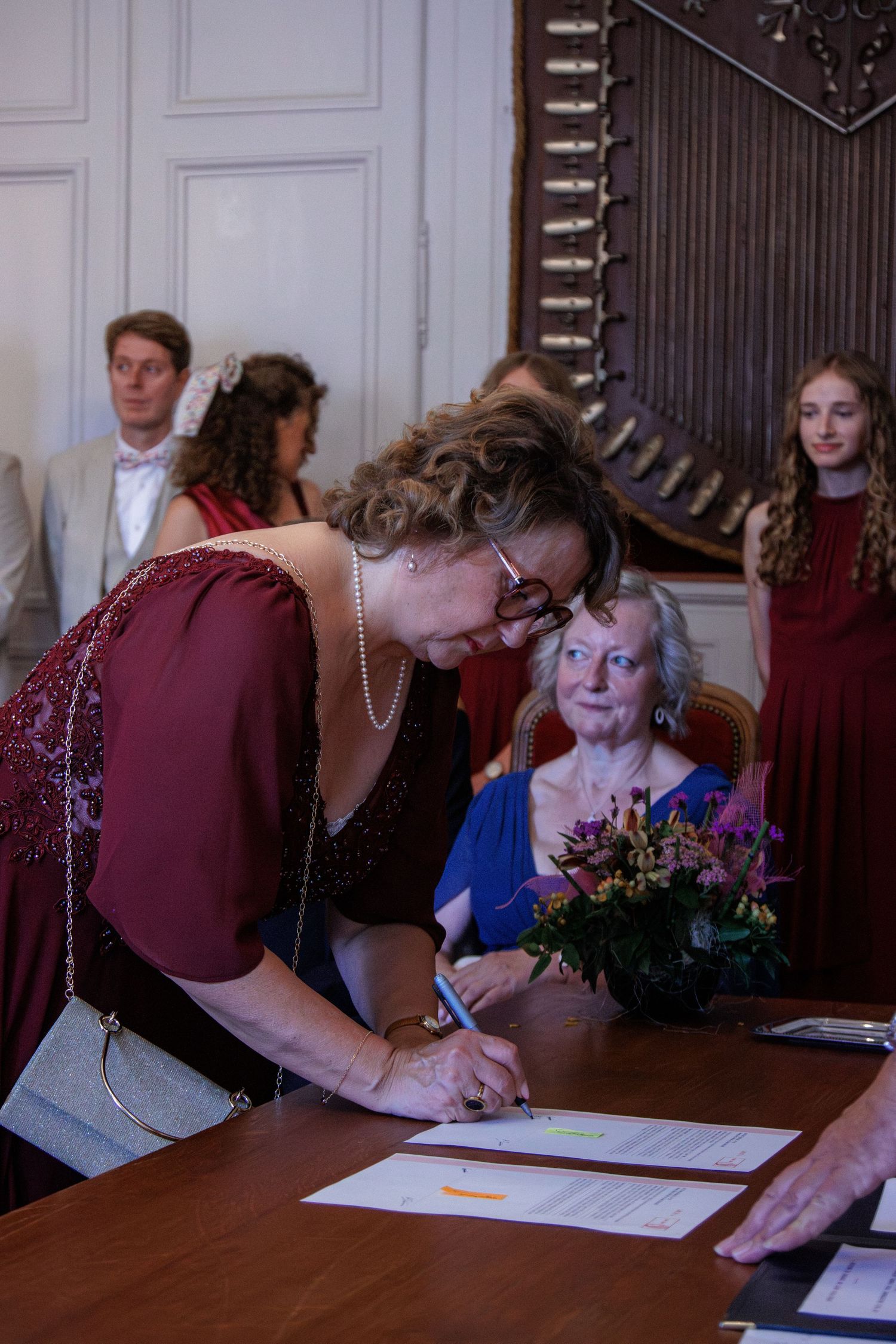 Woman in burgundy dress signing wedding documents at a table during a formal ceremony.
