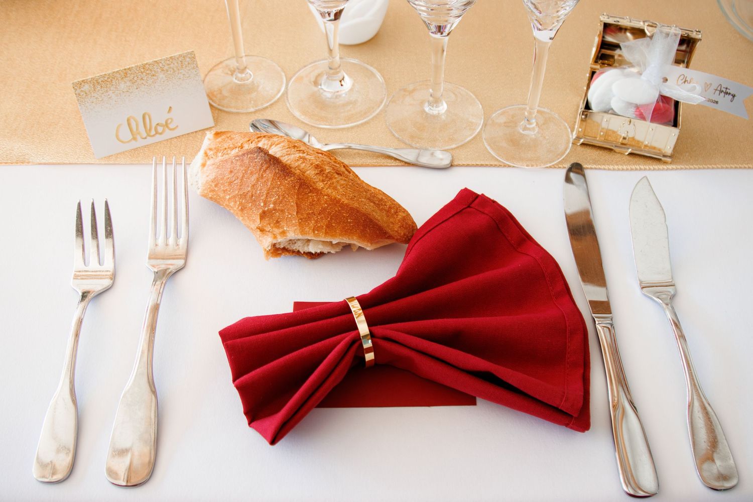 Elegant place setting with red folded napkin, silverware, wine glasses, and bread on a white tablecloth.