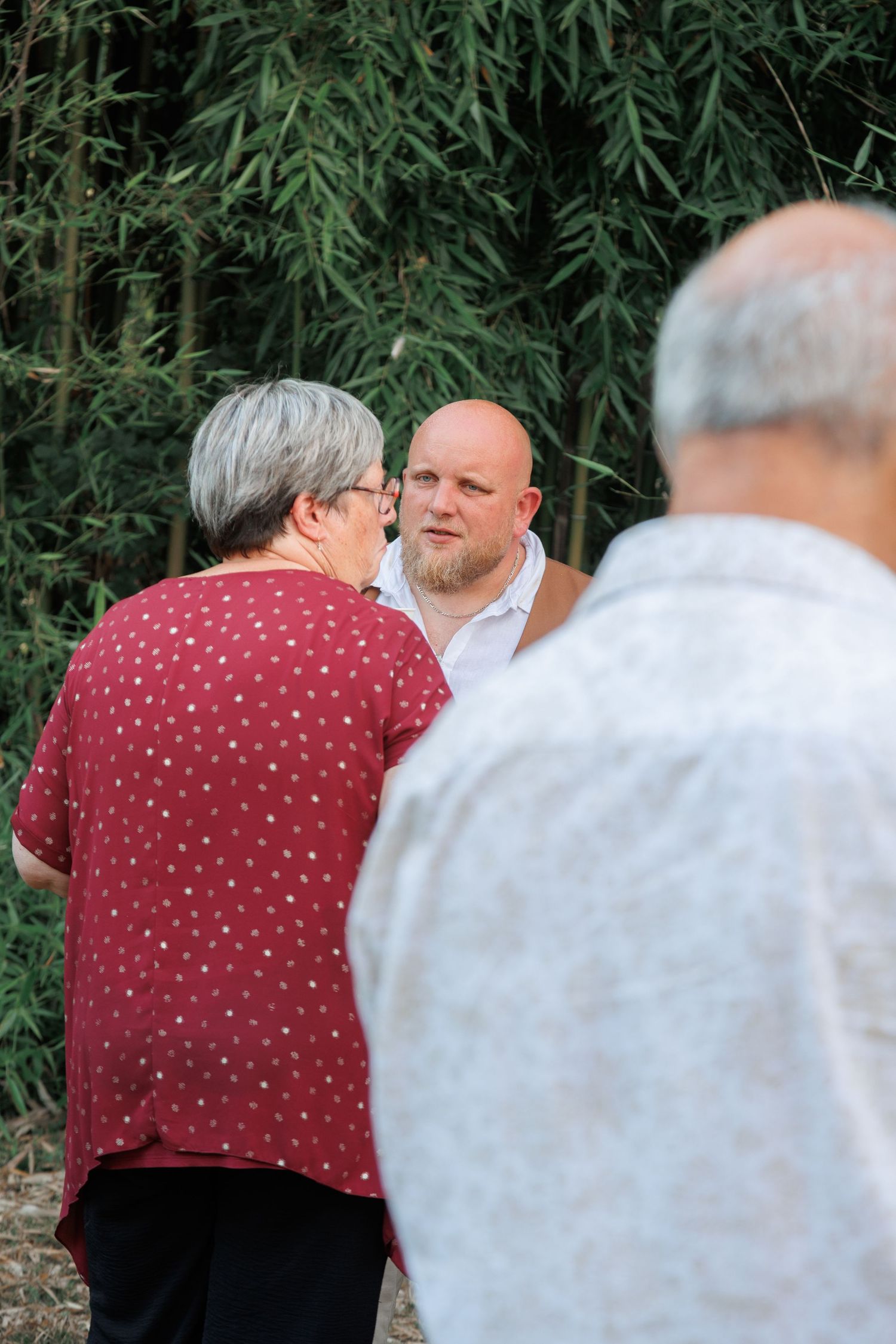 Bald man in white shirt engages in conversation with older woman in red polka dot dress outdoors.