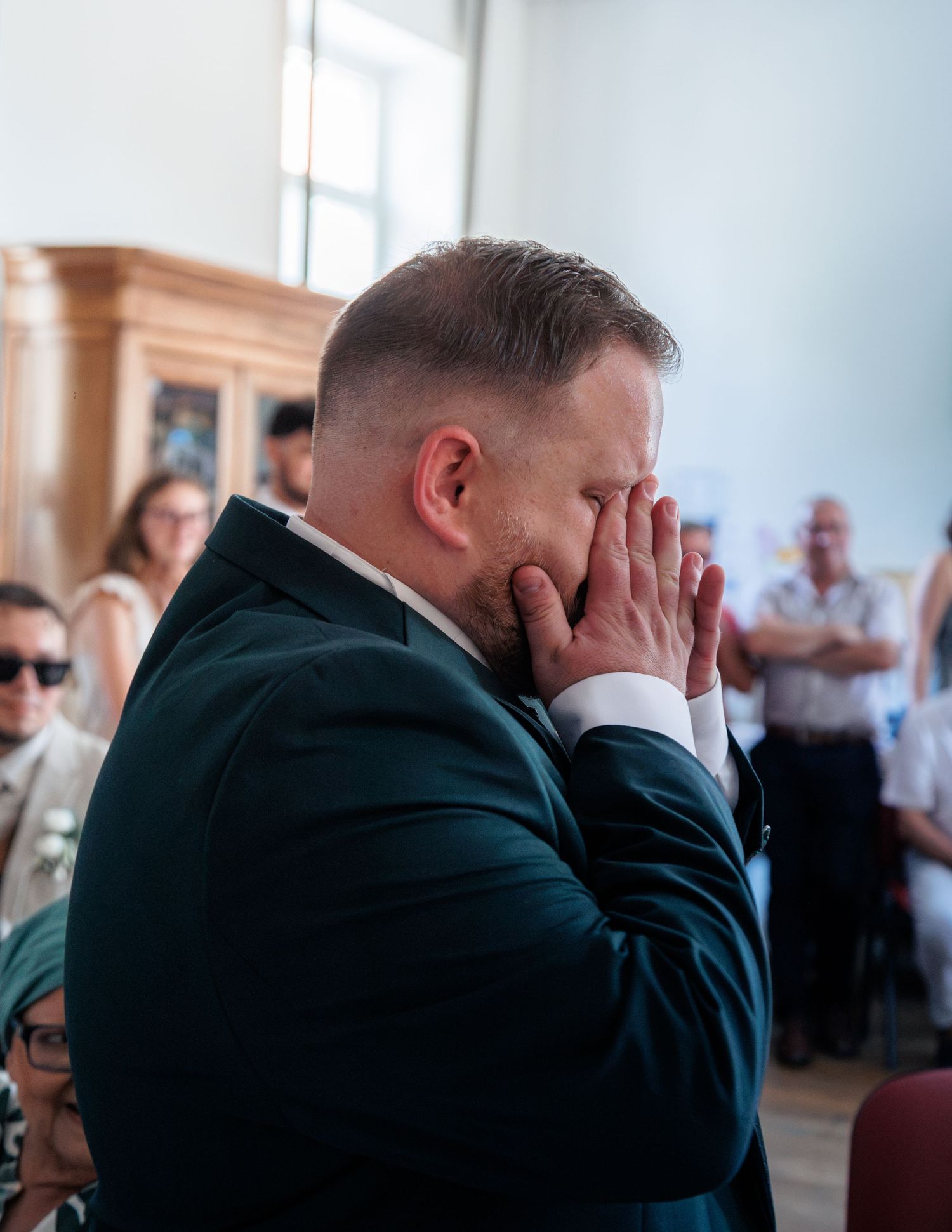 Emotional groom in teal suit covering face with hands during touching wedding ceremony moment.