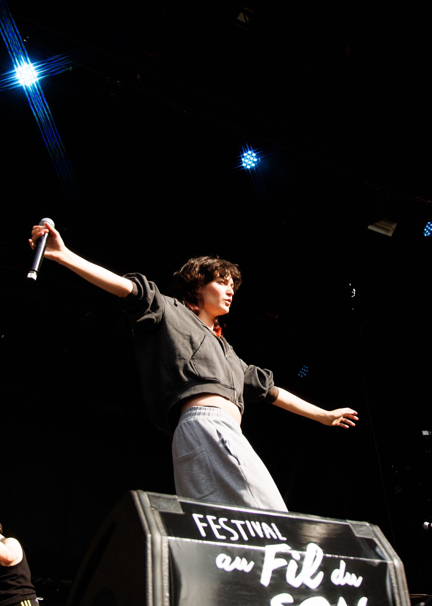 Performer on stage at Festival au Fil du Son, holding a microphone with arms outstretched under blue stage lights.