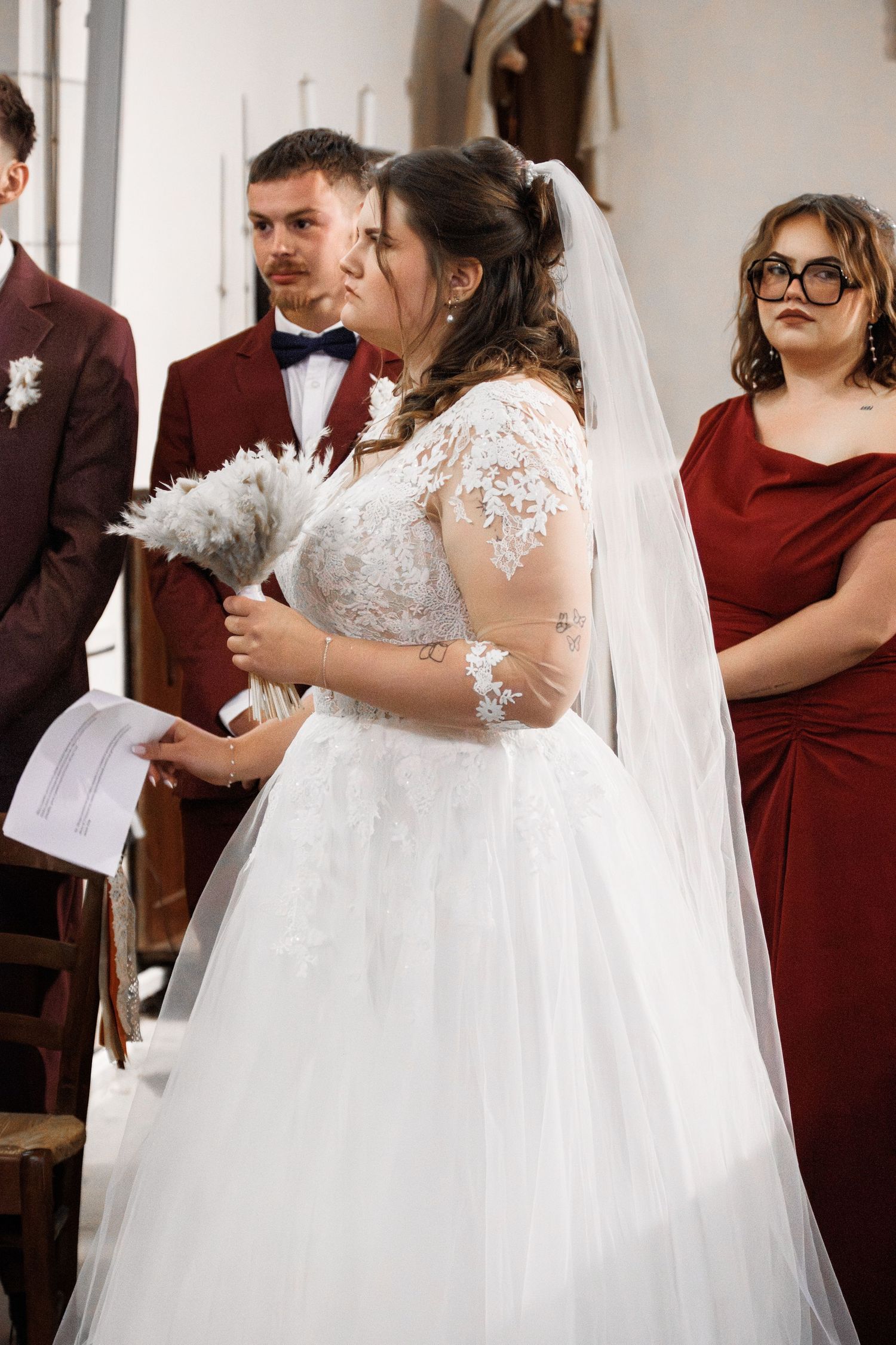 Bride in white lace gown and veil holds bouquet during wedding ceremony, with groom in burgundy suit behind her.