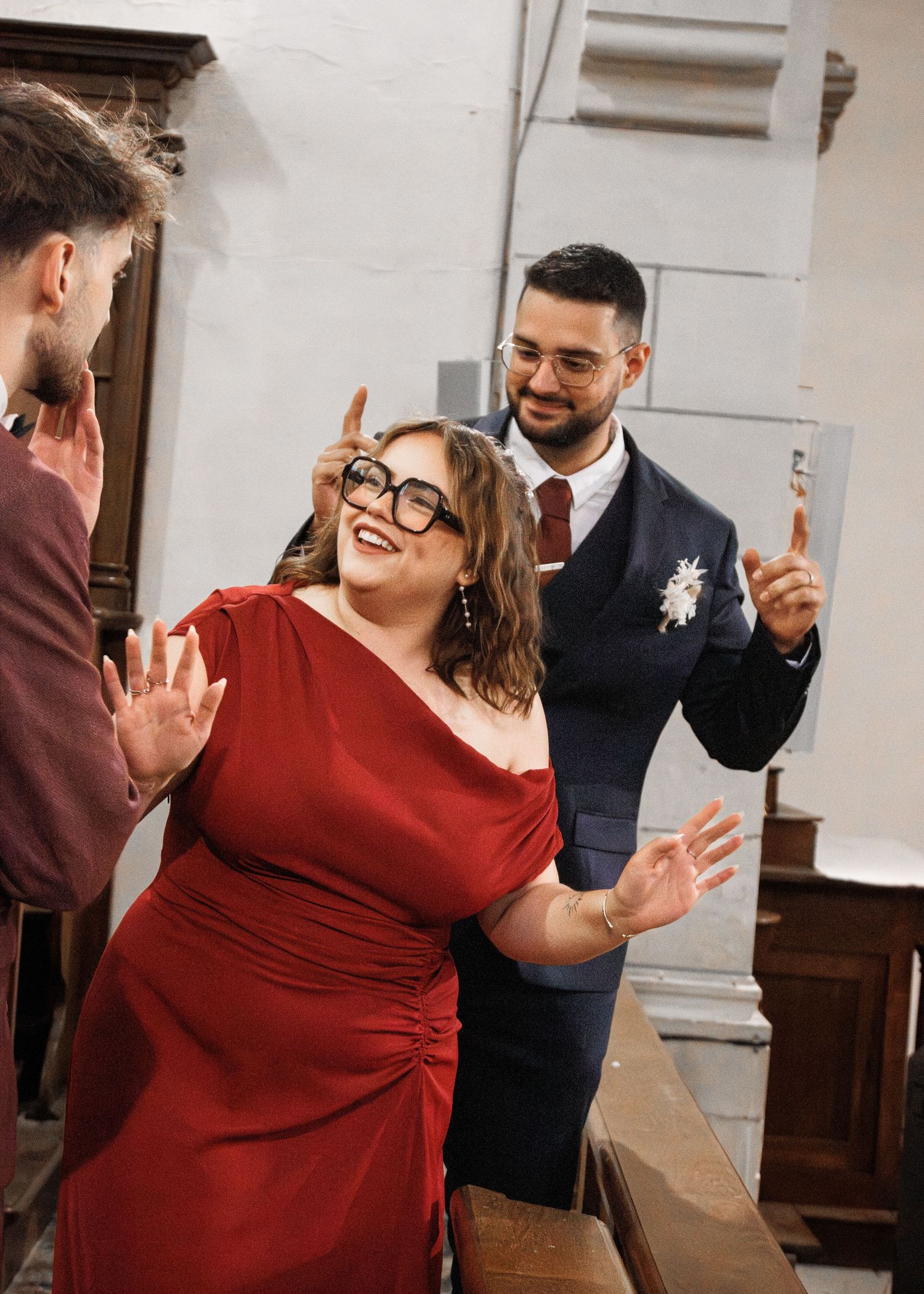Woman in red dress laughing while man in navy suit makes bunny ears behind her at a wedding celebration.