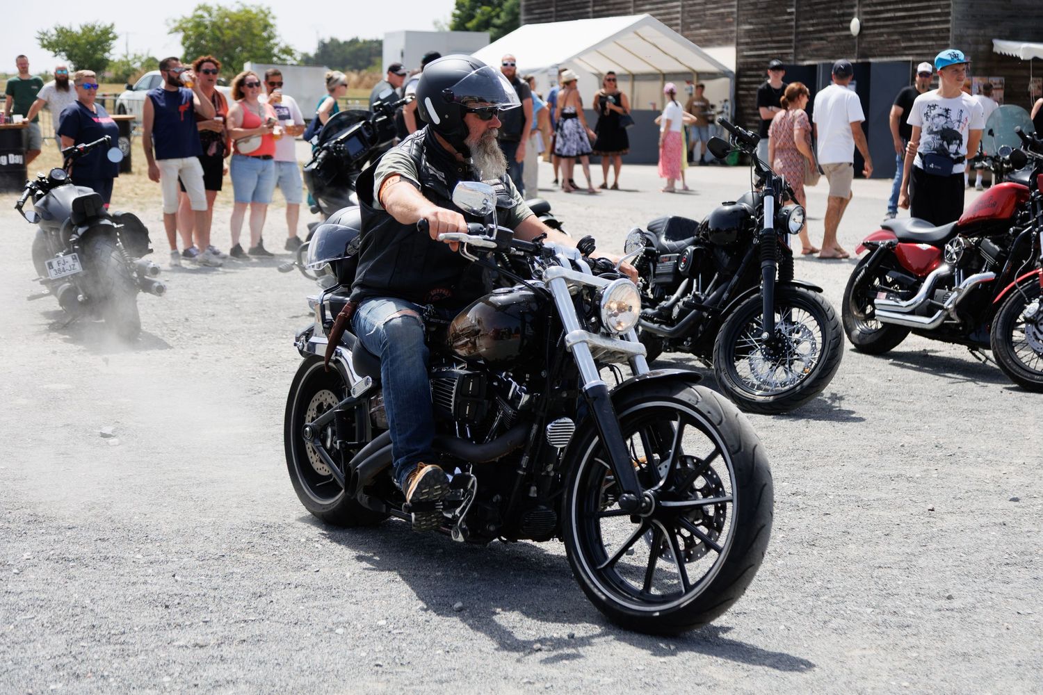 Motorcyclist riding a black Harley-Davidson at an outdoor biker rally event with spectators watching on a sunny day.