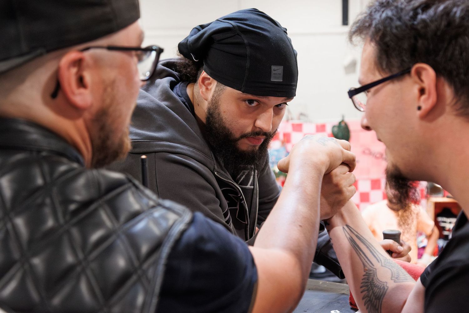 Three men with tattoos and casual attire engaged in an arm wrestling match at an indoor event.