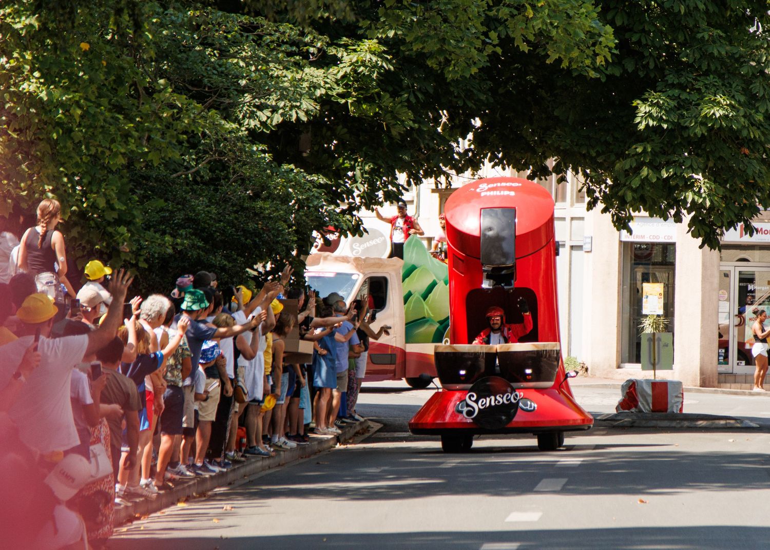 Spectators line the street watching a festive parade vehicle with red decorations pass by on a sunny day.