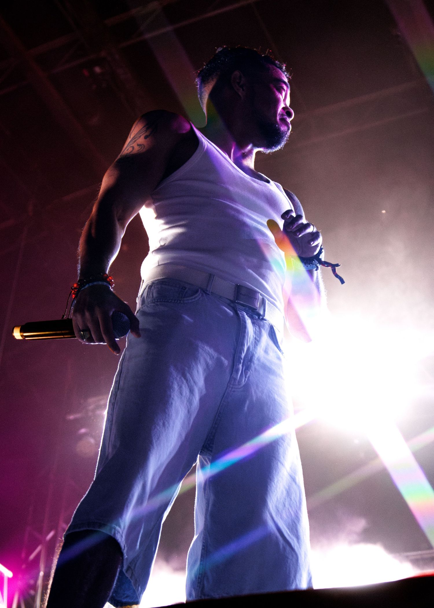 A performer in a white tank top and light-colored pants stands on stage bathed in dramatic purple lighting and lens flares.