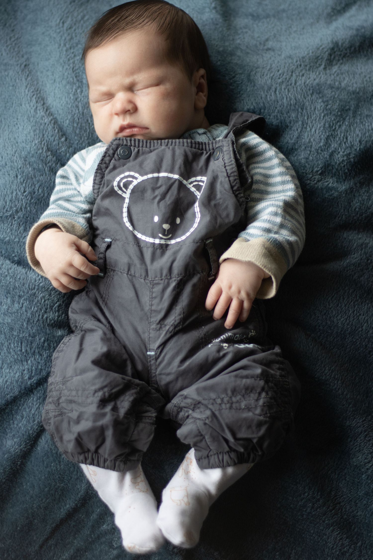 A baby sleeps peacefully wearing gray overalls with a bear design on dark blue fabric.