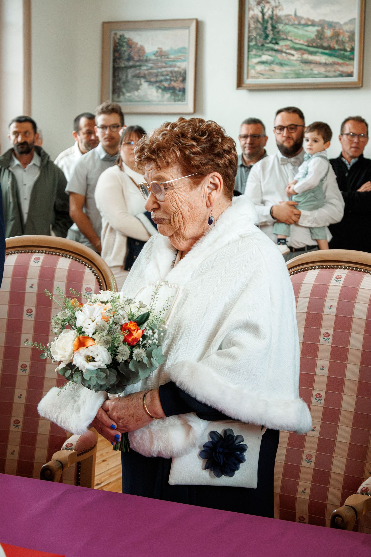 Elderly woman in white fur shawl holding a bouquet at a wedding ceremony, with guests standing in the background.