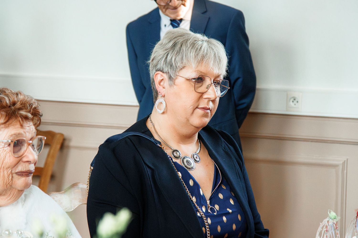 Elegant woman with short gray hair and glasses wearing a navy blazer and statement necklace at a formal event.