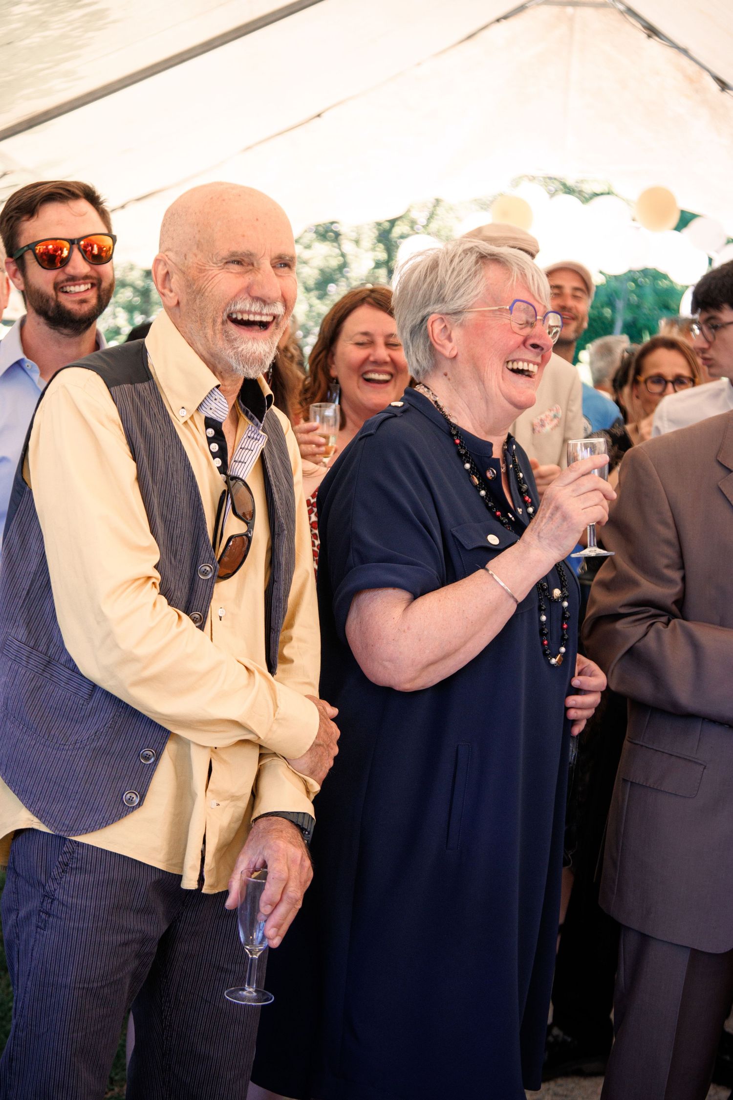 Joyful wedding guests laughing and celebrating under white tent at outdoor wedding reception.