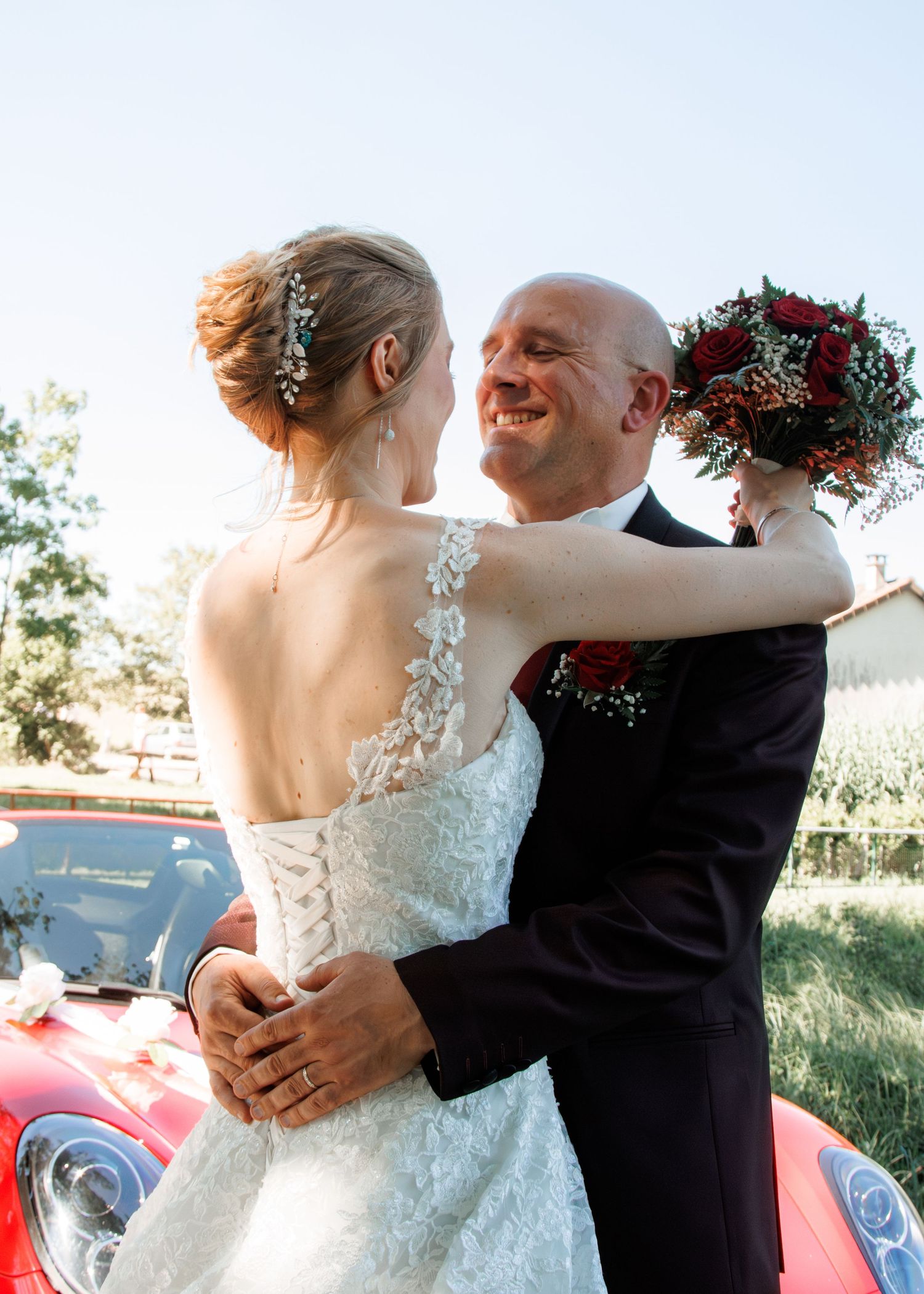 Groom in black suit embraces bride in lace wedding dress holding red roses bouquet near vintage red car.