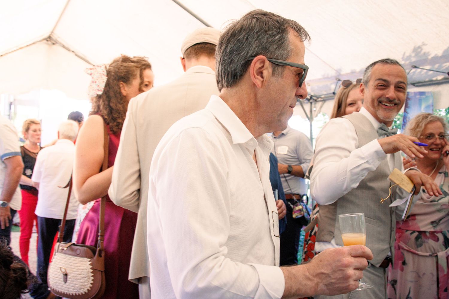 Man in white shirt holding a champagne glass mingling at a wedding reception under a tent.