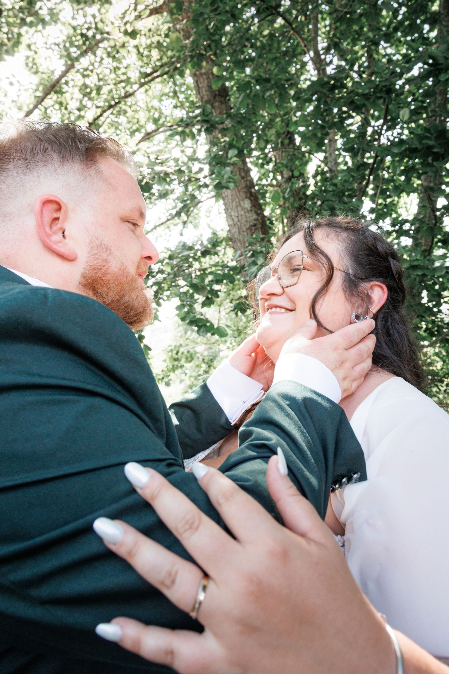 Groom tenderly cups brides face as she smiles joyfully under lush green trees after wedding ceremony.