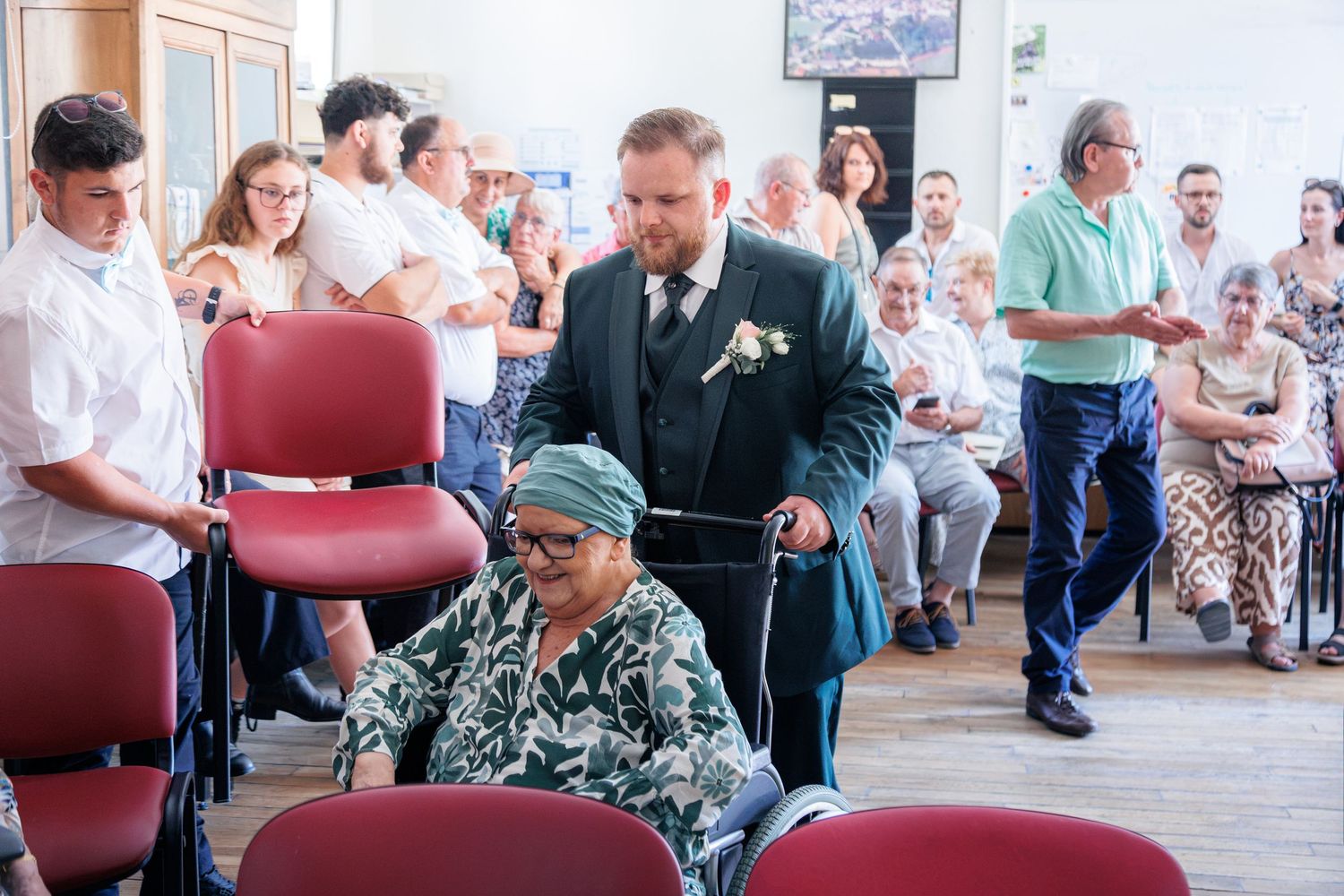 Groom in dark green suit escorts elderly woman in wheelchair through wedding ceremony venue.