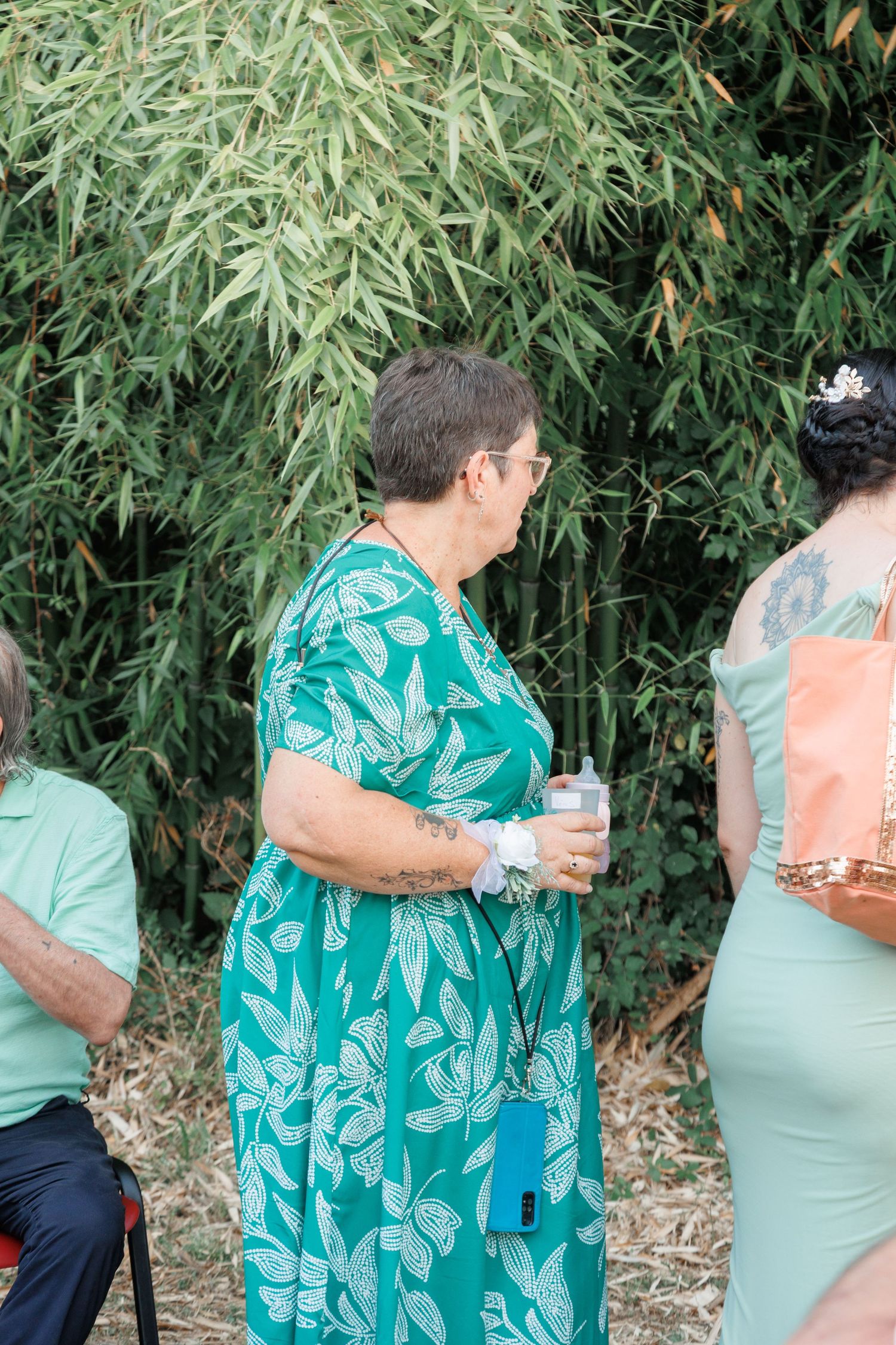 Wedding guest in teal floral dress holding drink outdoors near green bamboo foliage.