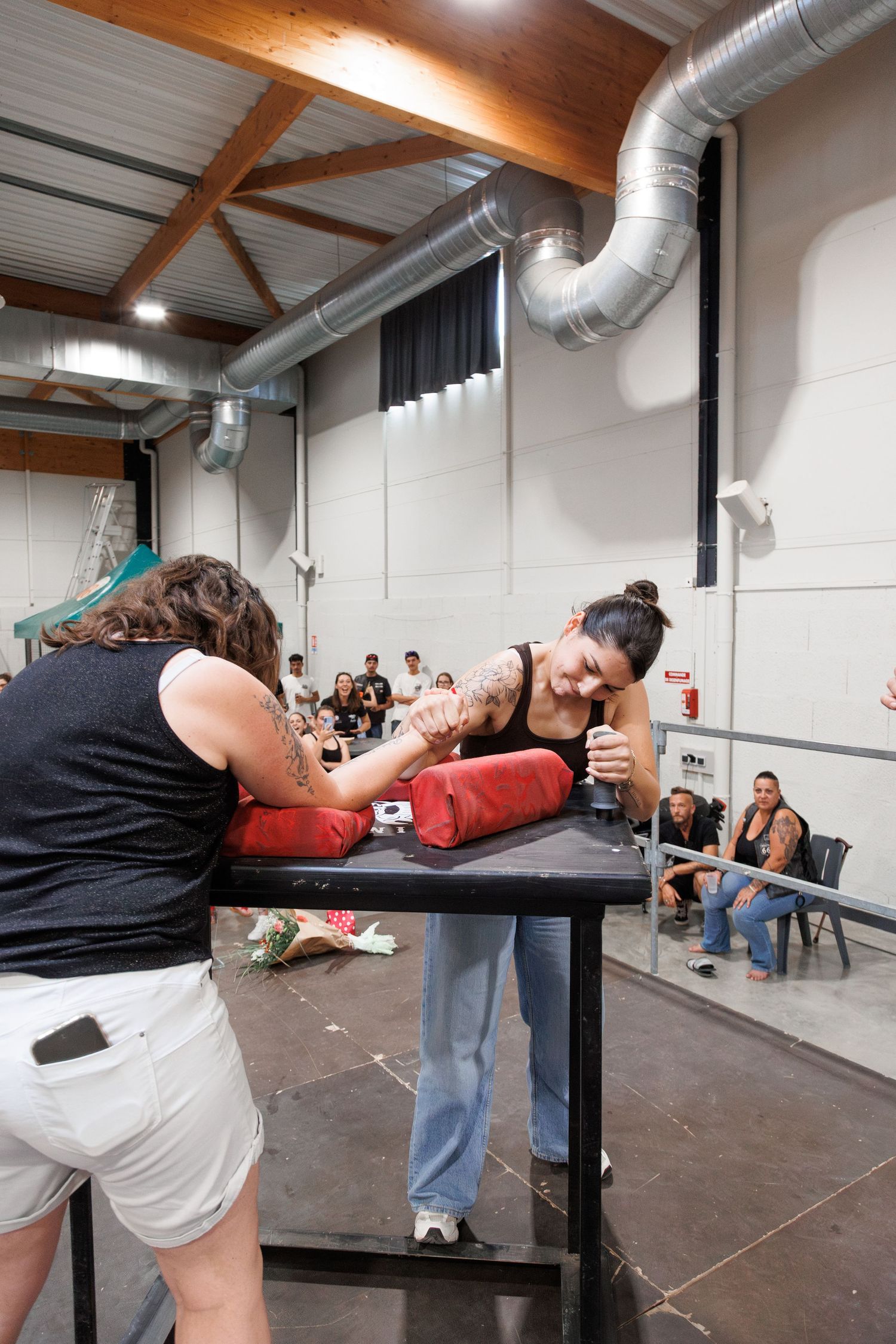 Two women arm wrestling on a padded table at an indoor competition event with spectators watching in the background.