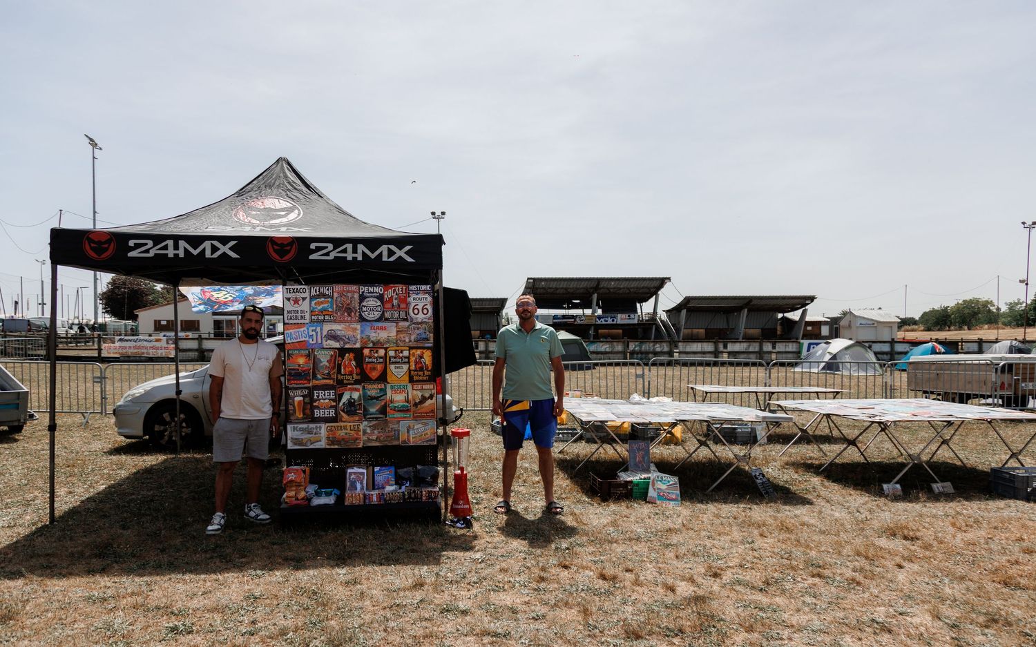 Two men stand at a 24MX branded merchandise tent booth at an outdoor motorsport event on a sunny day.