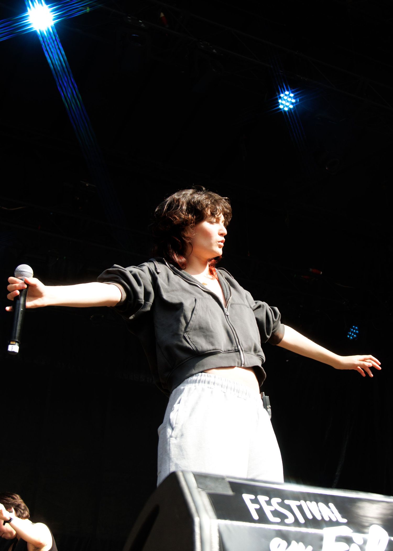 A performer holds a microphone on stage at a festival, arms outstretched, wearing a dark top and white skirt.