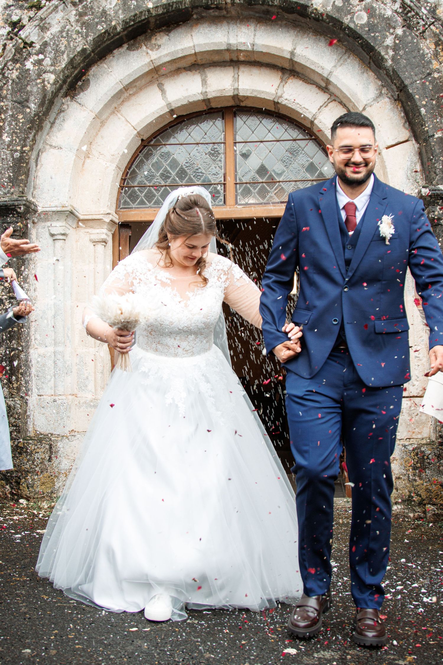 Newlywed couple exits stone church in confetti shower, bride in white gown, groom in navy suit, holding hands.