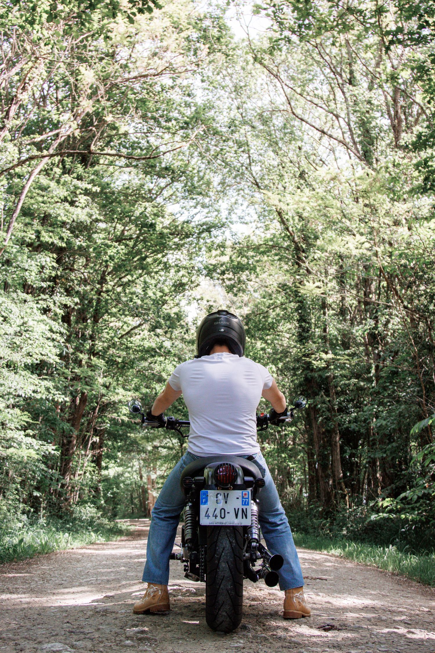 Person in white t-shirt and jeans sitting on motorcycle on forest dirt road, viewed from behind.