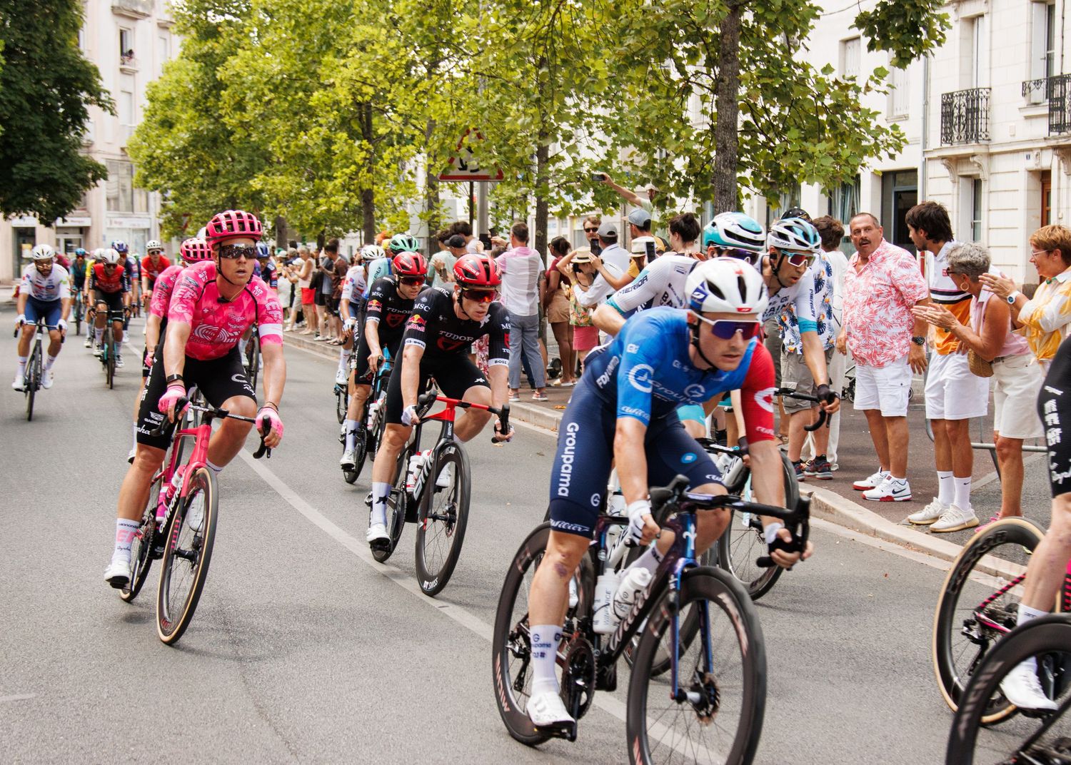 Cyclists in vibrant jerseys race through a city street while spectators cheer from behind barriers.