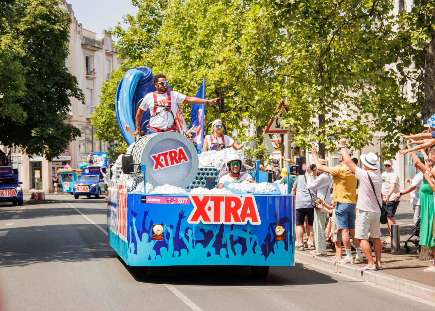 Colorful parade float with XTRA branding and blue decorative elements rolling down a tree-lined street.
