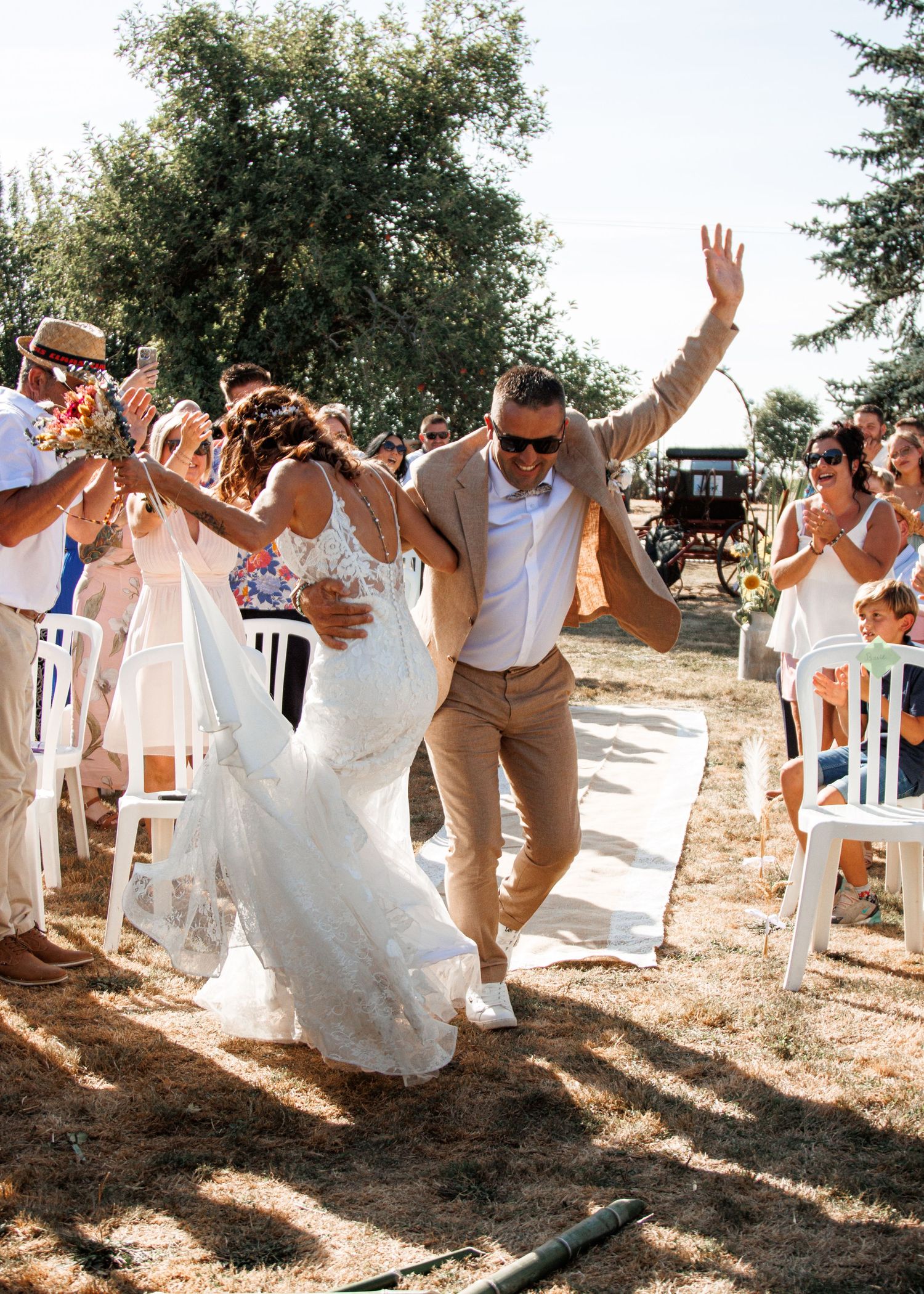 Wedding celebration with guests cheering and dancing on an outdoor path surrounded by trees on a sunny day.