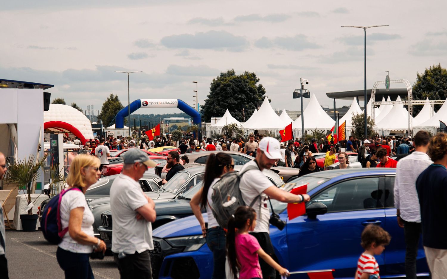 Crowd gathering at an outdoor car show with white tents and colorful vehicles on display.