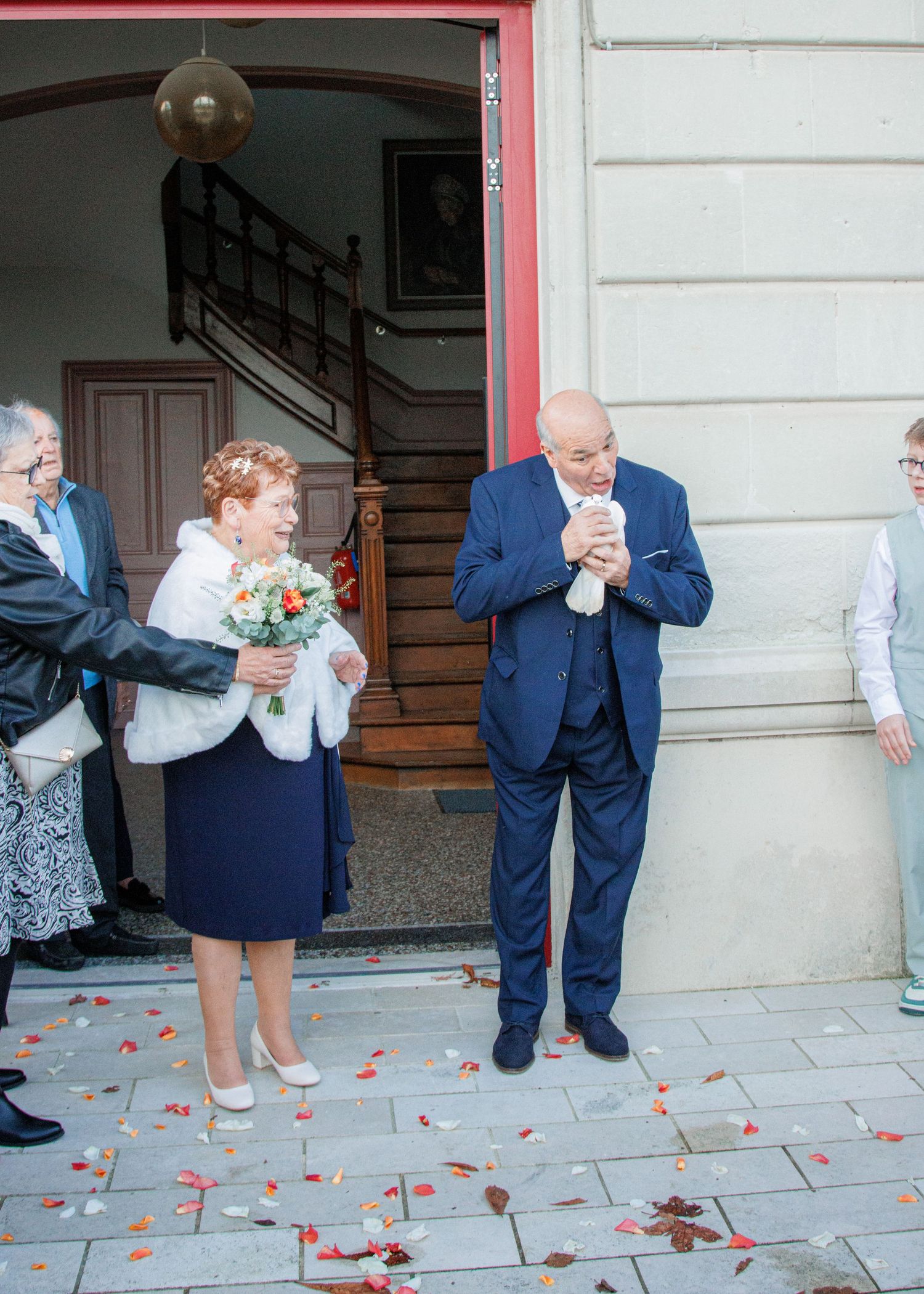 Elderly couple celebrating wedding outside building with flower petals, bride holding bouquet in white fur shawl and navy dress.