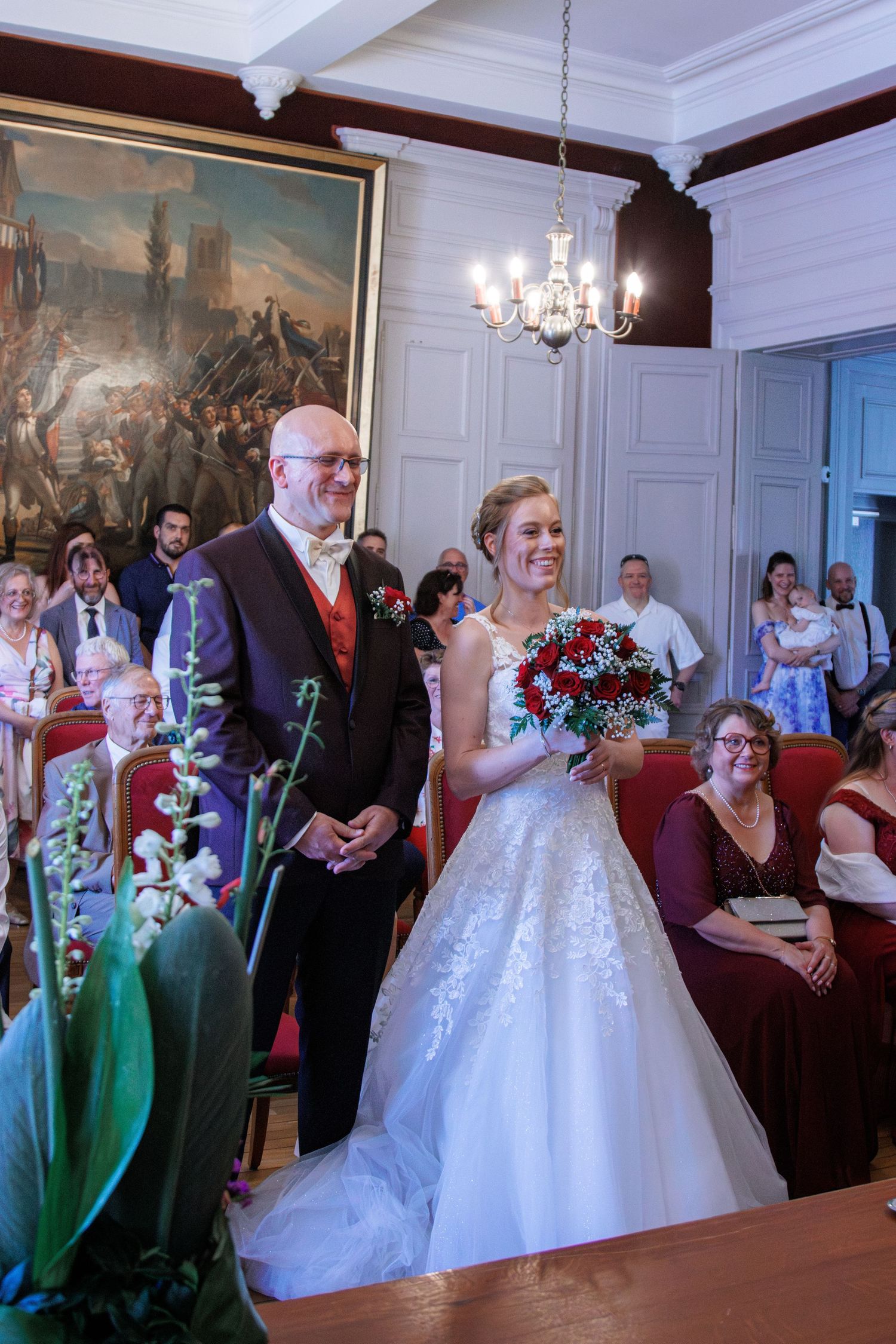 Smiling bride in lace gown holds red bouquet beside groom in dark suit at elegant wedding venue.