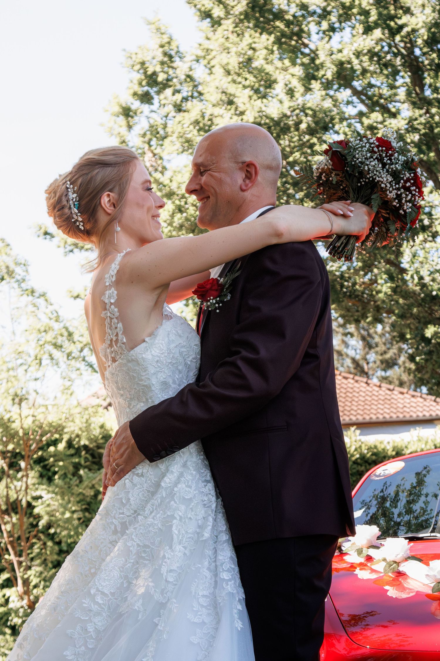 Bride in backless lace wedding dress embracing smiling bald groom outdoors near red car.