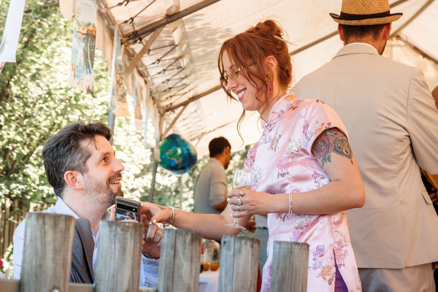 Man and woman in a floral dress laughing together at an outdoor wedding reception under a white tent.
