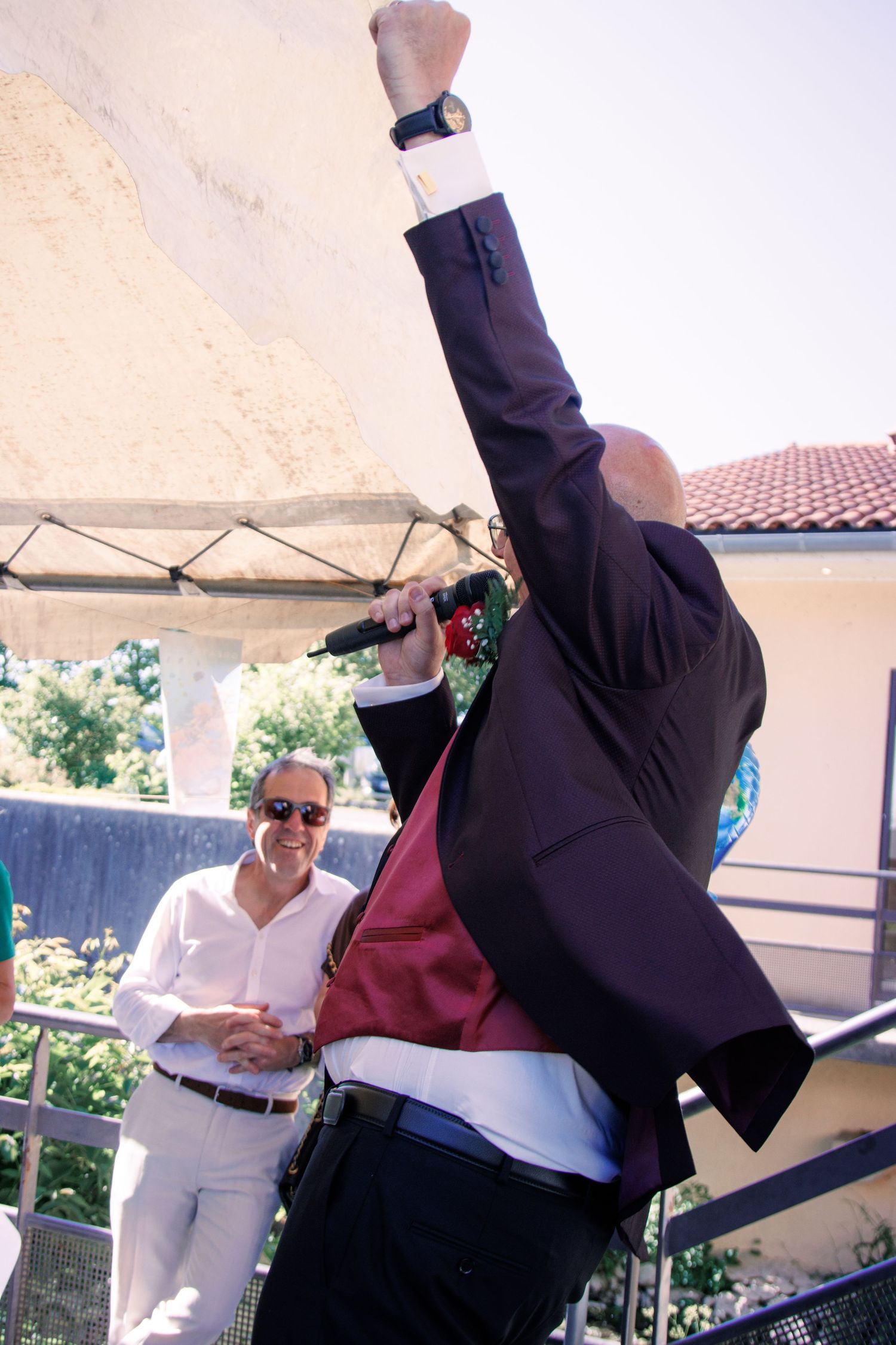 Energetic performer raises fist holding microphone on outdoor stage while smiling man watches at wedding celebration.