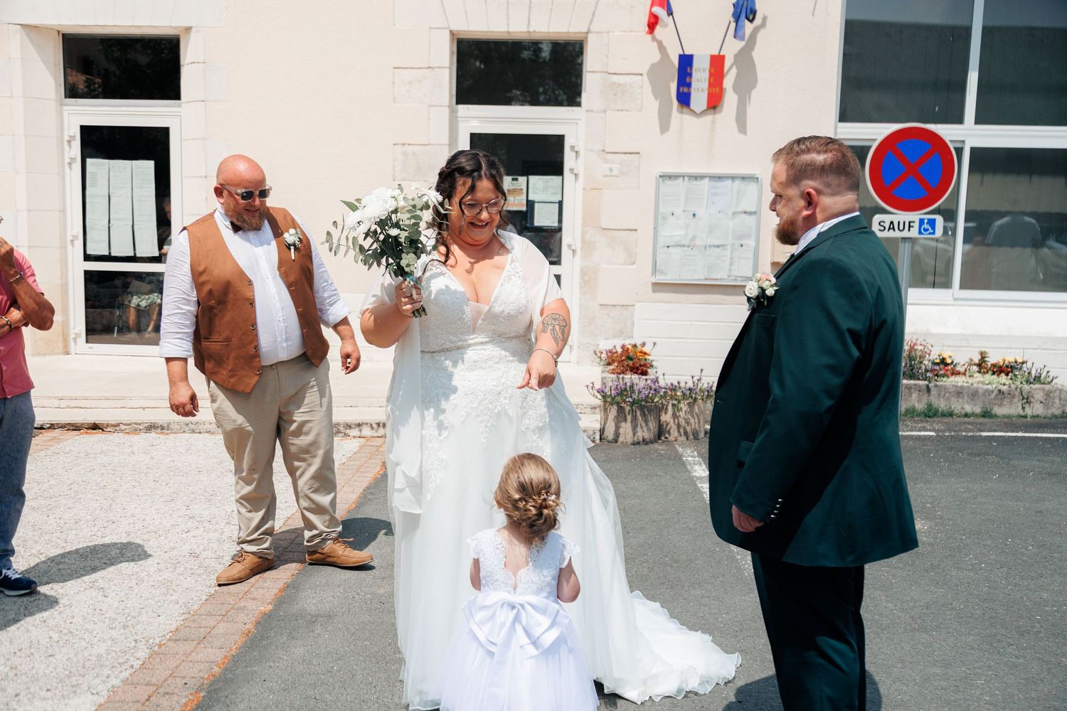 Bride in white gown holds bouquet while greeting groom outside French mairie with flower girl.