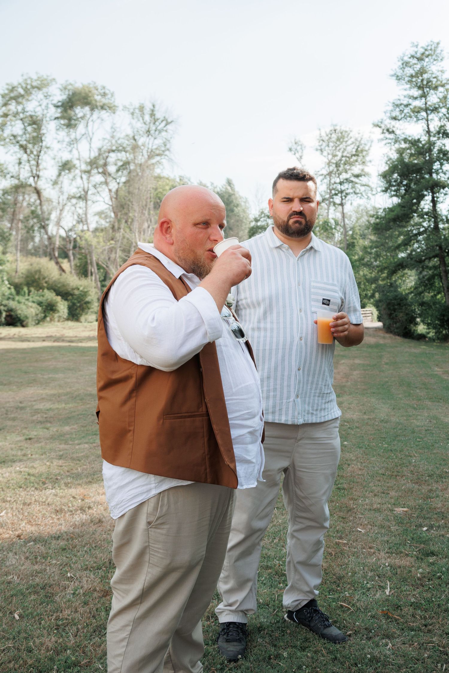 Two male wedding guests enjoying drinks outdoors in a green park setting.