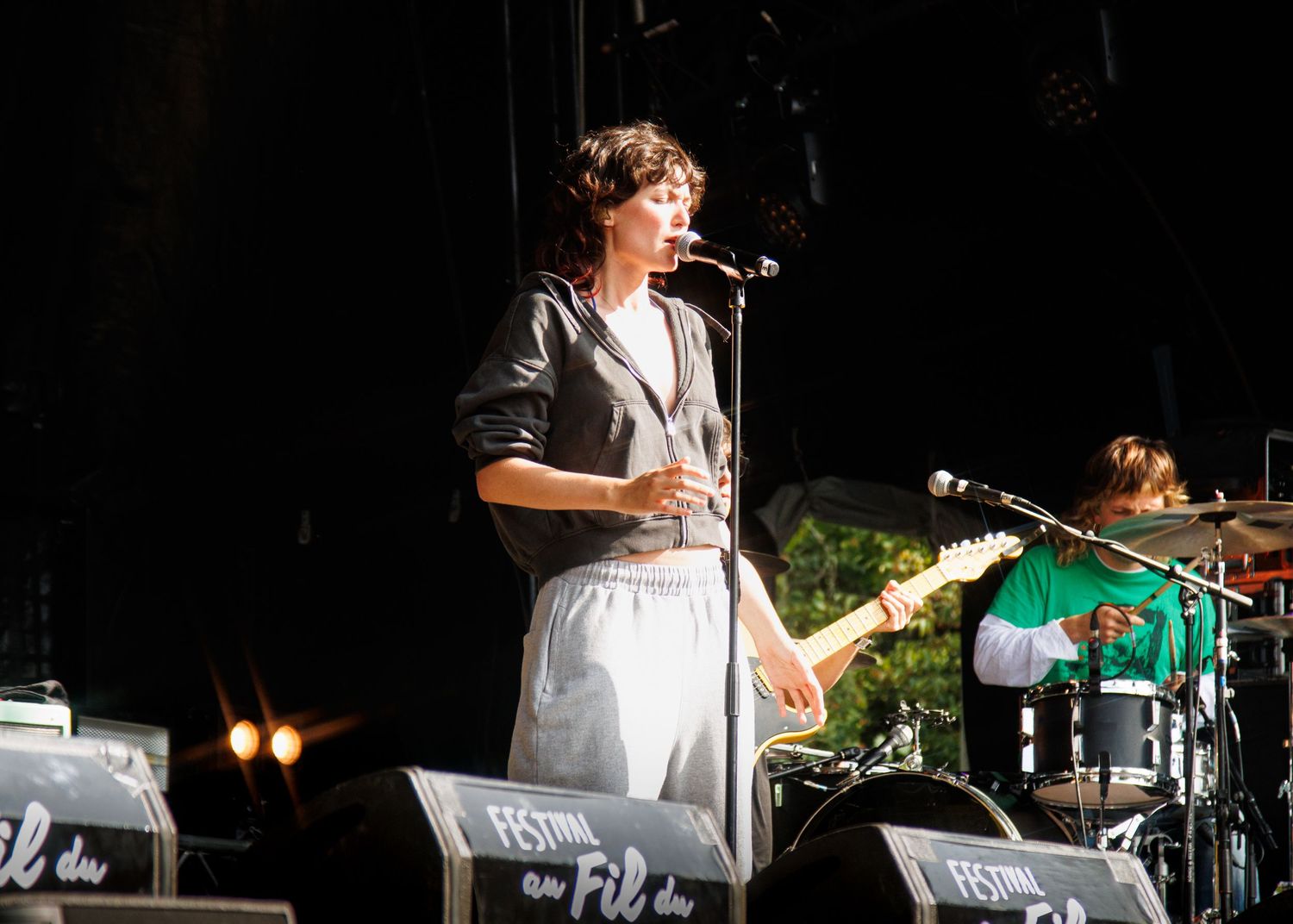 Singer performing on outdoor festival stage with guitarist in green shirt and drums visible in background.