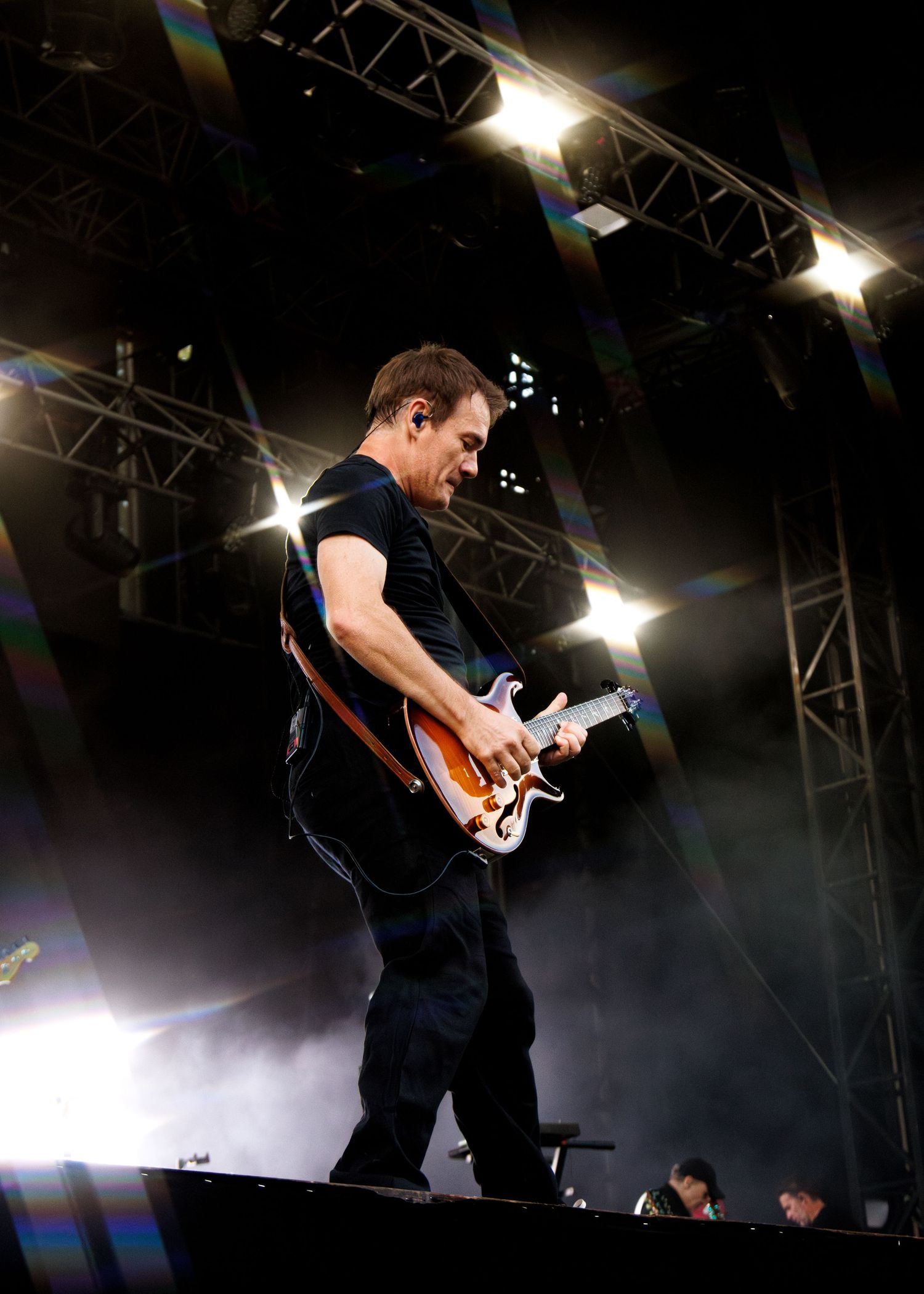 Guitarist performing on stage under bright lights at an outdoor concert, playing an electric guitar in a black t-shirt.