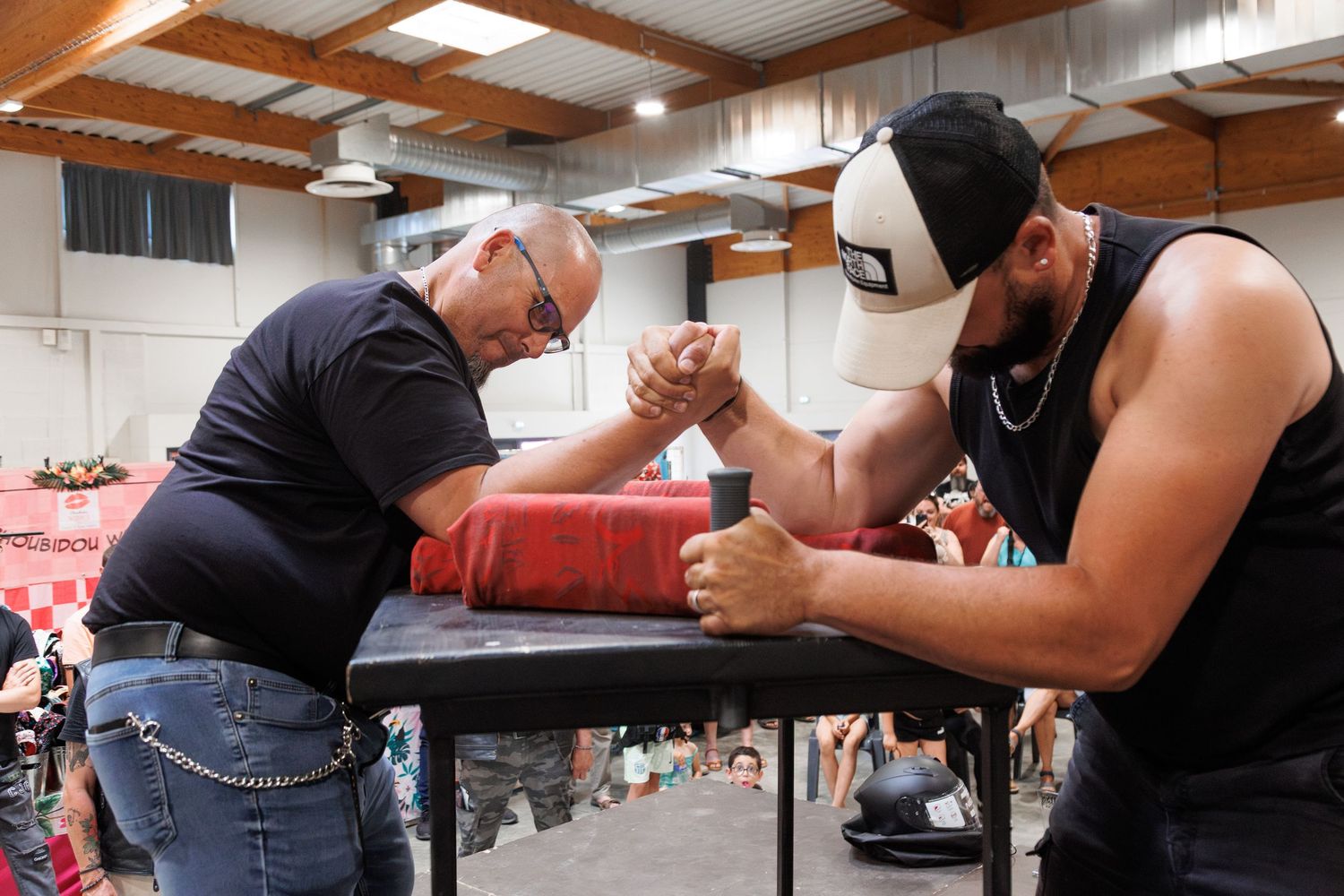 Two men competing in an arm wrestling match at an indoor tournament, leaning over a black table.