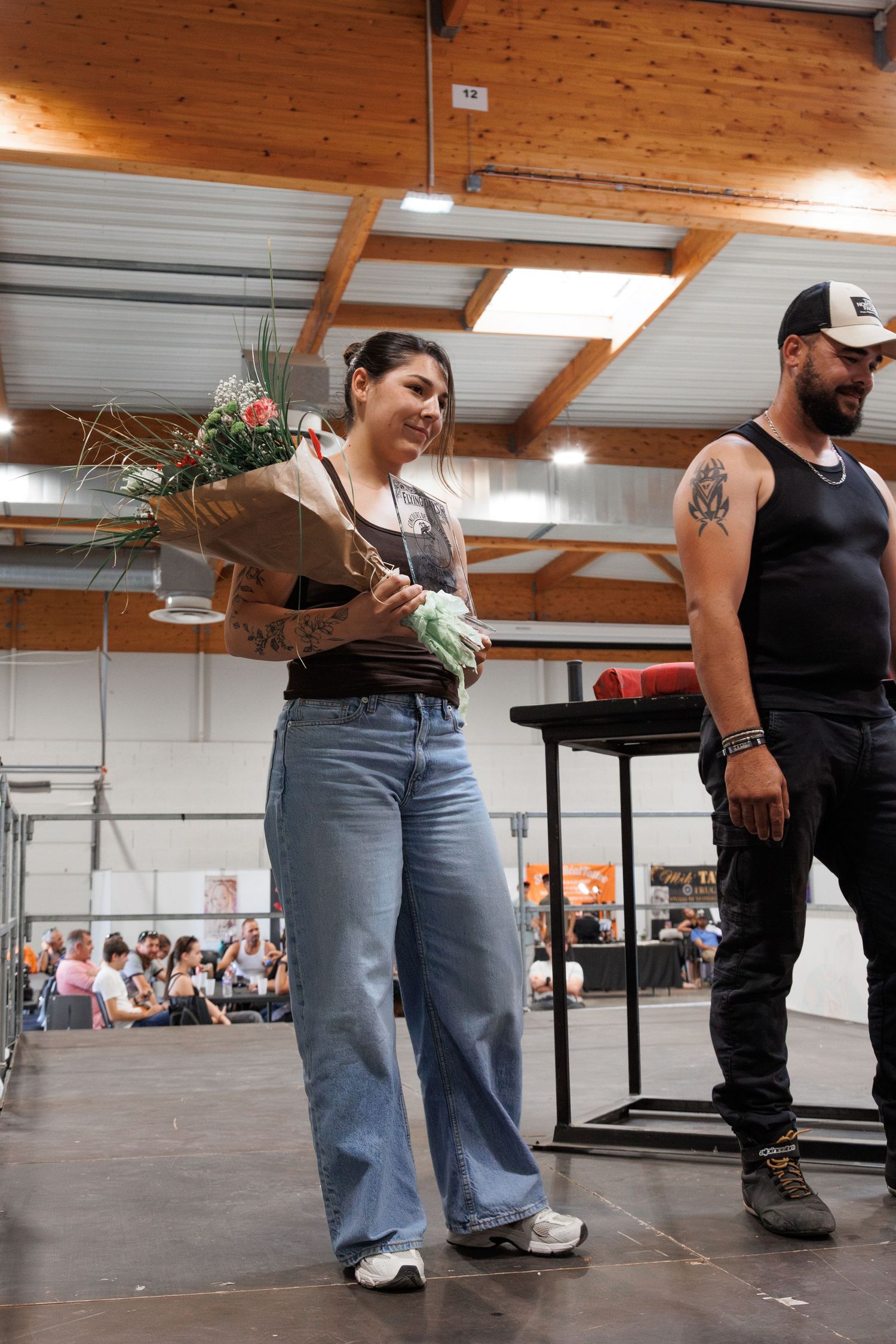 Woman holding flower bouquet on stage at indoor event, smiling beside tattooed man in black tank top.