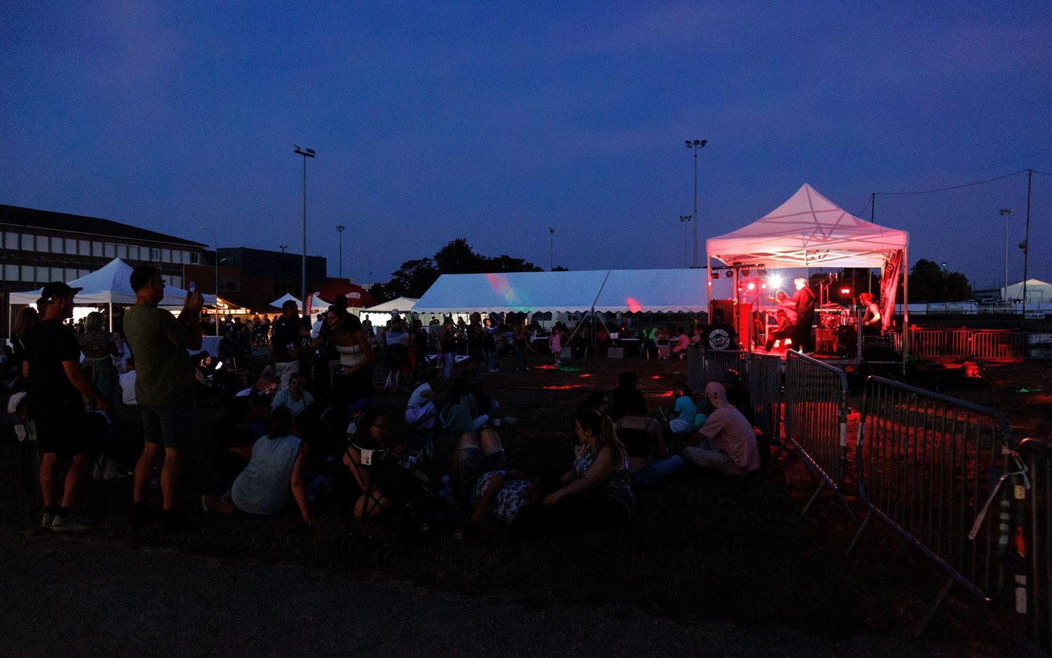 Outdoor evening concert with crowd gathered near illuminated stage tent at dusk.