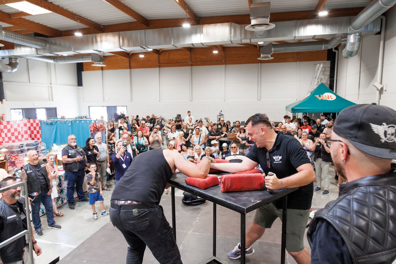 Two competitors arm wrestling at a packed indoor event, watched by a large crowd of spectators.