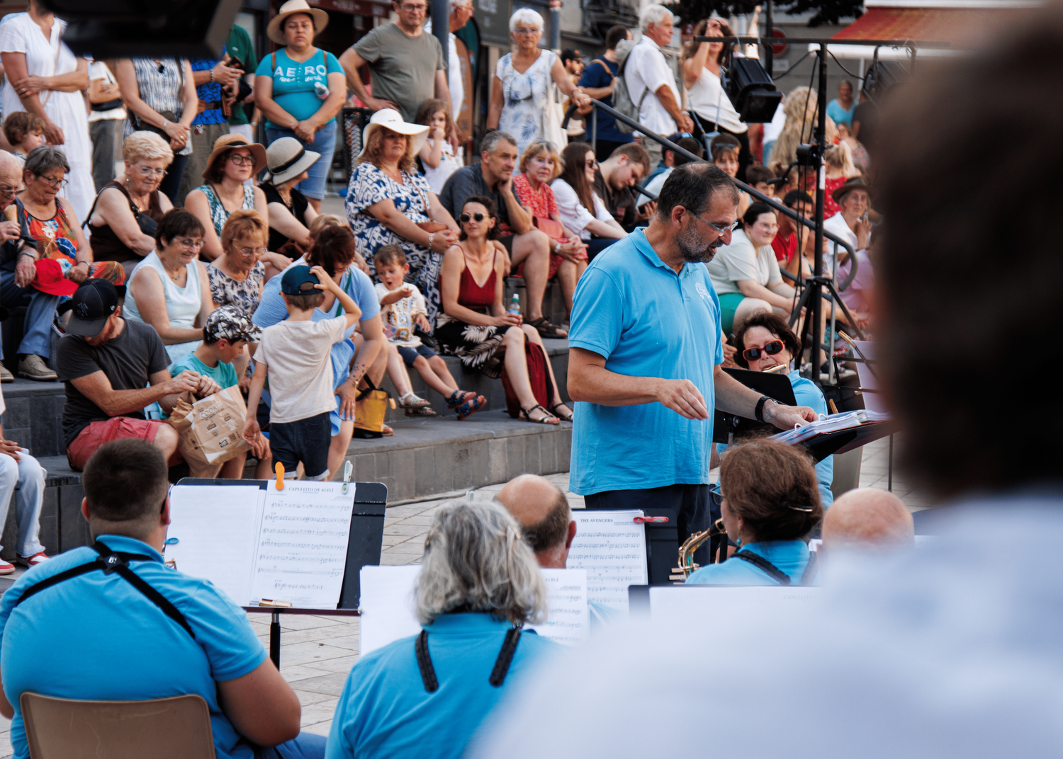 A conductor leads an outdoor band performance while crowd watches from stadium seating.