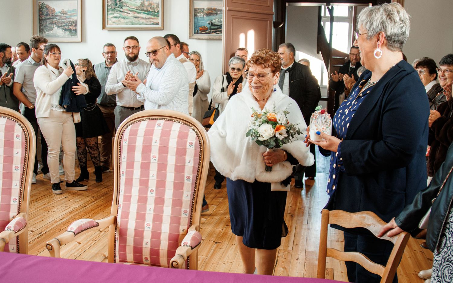 Bride holding bouquet in white fur shawl greets guests at elegant indoor wedding ceremony.
