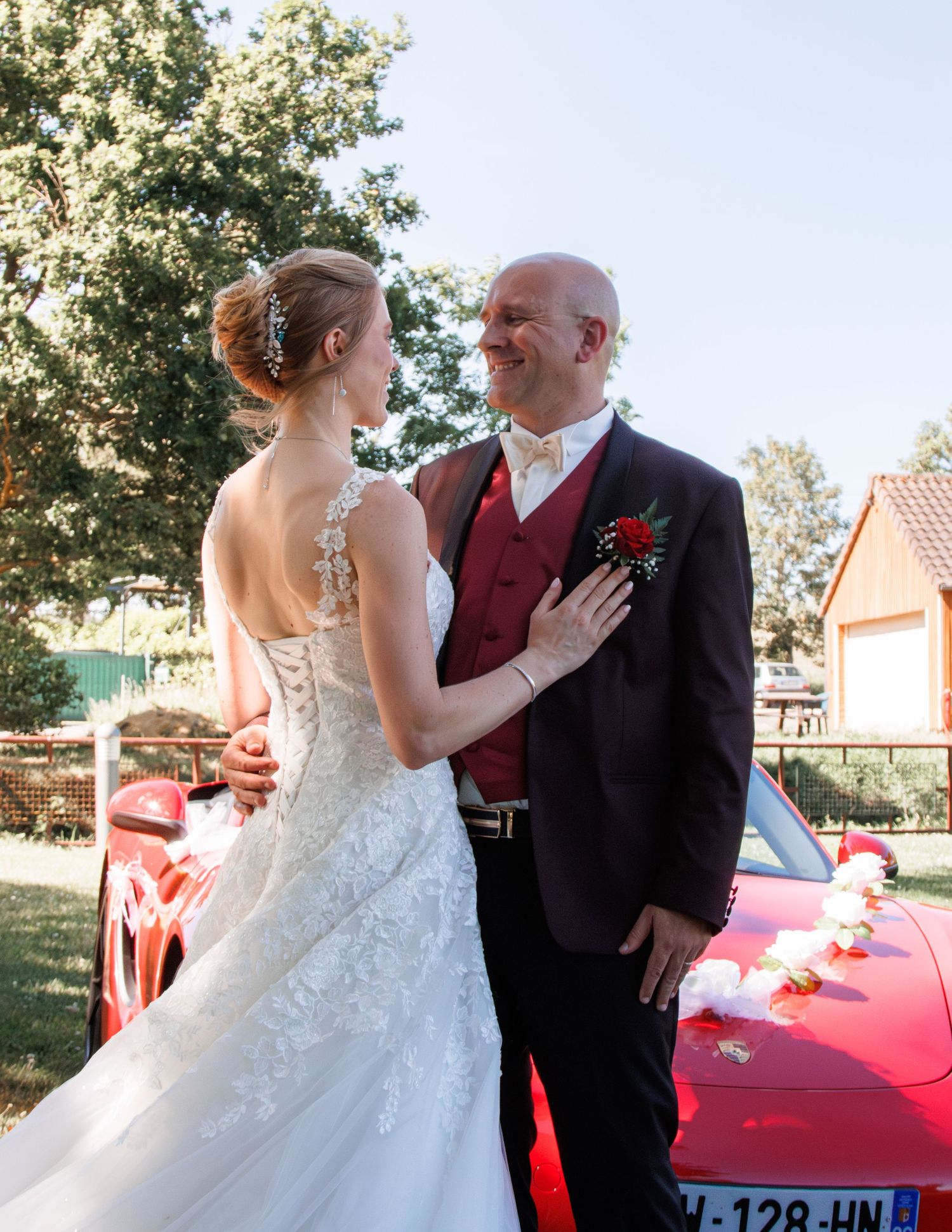 Bride in lace gown and groom in dark suit with red vest share romantic moment by red car.