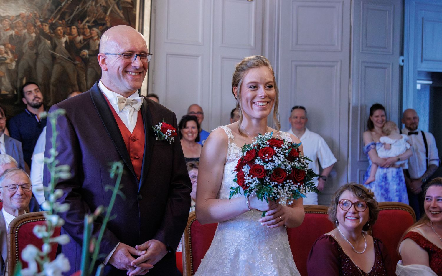 Bride holding red and white bouquet walks down aisle with bald groom in red vest at wedding ceremony.