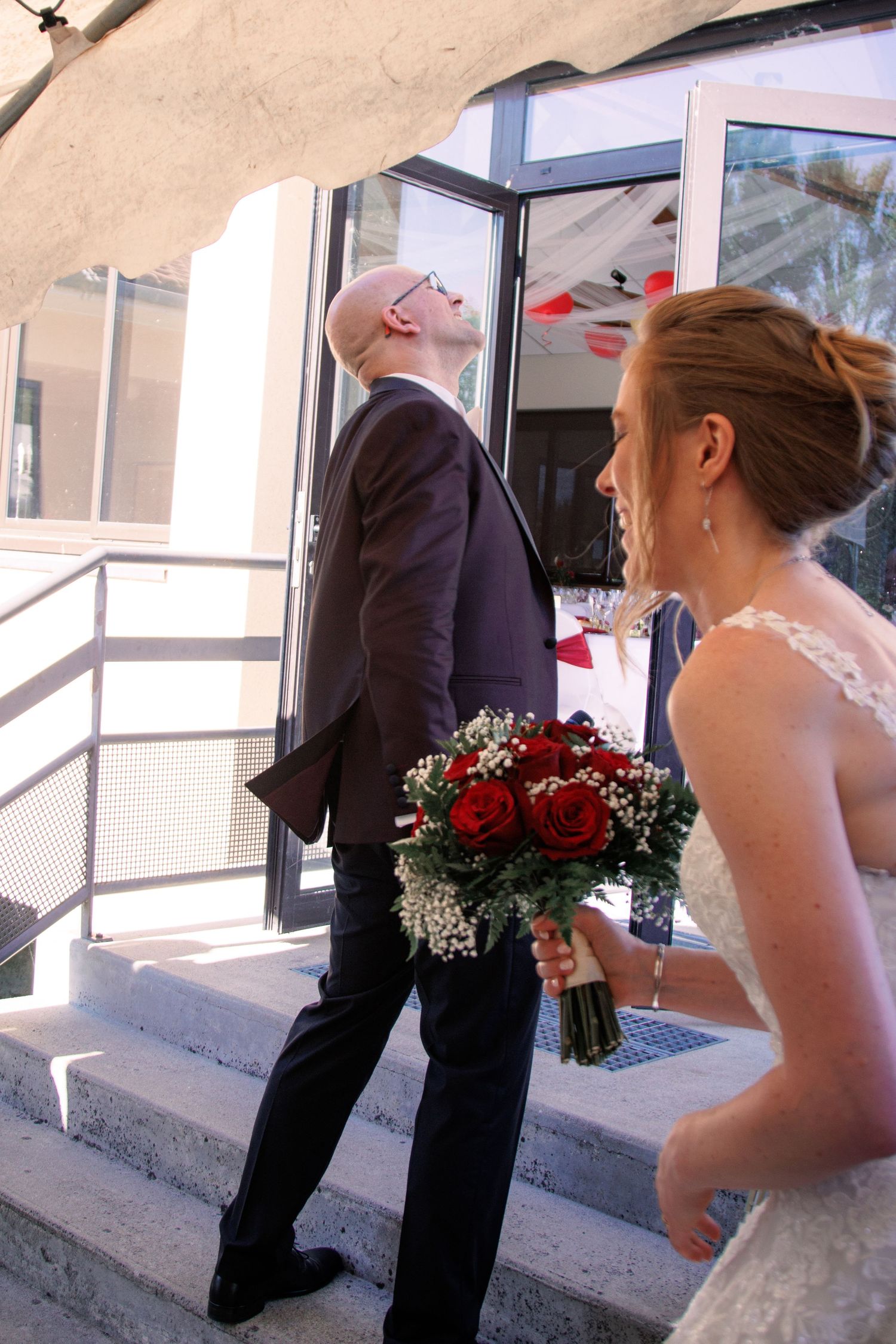 Bald groom in dark suit looks up at building entrance while bride holds red rose bouquet on steps.