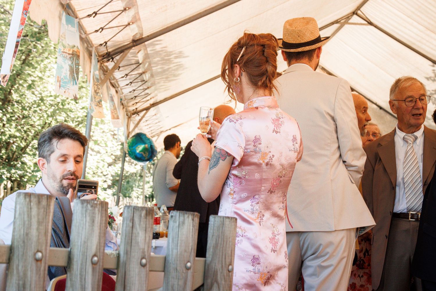 Wedding guests socializing under a sunny outdoor tent, woman in floral dress holding a champagne glass.